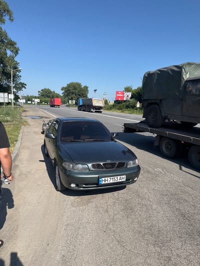 On a sunny day, on the side of a rural road in Prybuzke, Ukraine, a dark green Daewoo Nubira sedan is parked. The car is facing forward with its front passenger side towards the camera. The license plate reads "HH 7153 AH". The car appears to be connected to a trailer, which is also visible. In the background, a military-style vehicle is being transported on a flatbed truck. Further down the road, a red truck and a blue dump truck are visible. The environment is characterized by a clear blue sky, trees on the sides of the road, and utility poles. A person in a black t-shirt and jeans is standing to the left of the frame, holding a can.