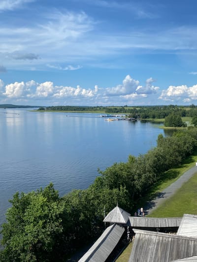 The image captures a scenic view of a wide, tranquil lake on a bright, sunny day in Boyarshchina, Russia. The sky is a vivid blue, dotted with fluffy white clouds. The lake, reflecting the sky, stretches towards a distant tree-lined shore. Several small boats and a dock with moored vessels are visible on the water. In the foreground, a pathway winds through a grassy area bordered by lush green trees. Wooden structures, possibly part of an architectural ensemble, with characteristic roofs and wooden walkways, are prominent in the lower right portion of the image. Two individuals, a child and two adults, are walking along the pathway. The overall impression is one of a peaceful, natural setting with elements of human activity and historical architecture.