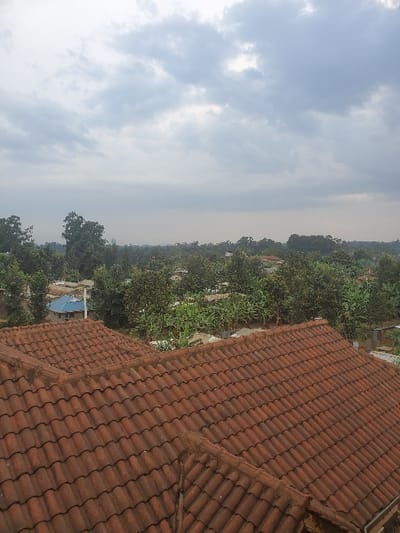 An elevated wide-angle view depicts a residential landscape in Kihara ward, Kenya, under an overcast sky. The foreground is dominated by multiple pitched sections of a reddish-brown tiled roof, exhibiting curved, interlocking tiles with signs of weathering and minor debris accumulation. Beyond this structure, the midground presents a dense canopy of varying green trees and vegetation, interspersed with numerous residential buildings. A prominent light blue corrugated metal roof is visible on a building to the left, while other structures feature gray or earth-toned roofs. The background consists of a distant tree line and a hazy horizon, beneath a sky uniformly covered with gray and white clouds. No individuals, specific actions, or interactions are discernible within the frame.