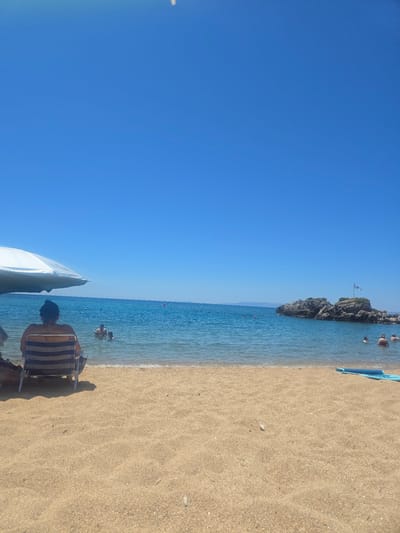 A wide shot captures a serene beach scene on a clear, sunny day in the Municipal Unit of Lefktro, Greece. The foreground is dominated by golden sand, with a blue inflatable mat resting on the right. In the mid-ground, gentle waves lap the shore, with several people swimming and wading in the turquoise water. To the left, a white beach umbrella provides shade for a person sitting in a striped beach chair, their back to the camera. Further out in the sea, a rocky islet is visible, topped with a flagstaff flying a white flag. The background features a vast, clear blue sky meeting the horizon, with faint landmasses visible in the distance. The overall atmosphere is one of peaceful relaxation and summer enjoyment.