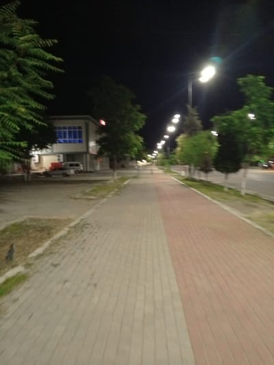 The media file depicts an empty street scene at night in Dustlik, Uzbekistan. The foreground is dominated by a paved walkway, with one section made of light-colored rectangular pavers and an adjacent section made of reddish-brown pavers. To the left of the walkway is a strip of dry grass and some dirt.
Beyond the walkway, a road is visible, lined with streetlights that illuminate the scene. Trees with green foliage are planted along the side of the road, some appearing tall and slender, others fuller. In the background, a building with a modern facade, featuring large blue-tinted windows, stands illuminated. A car, possibly a light-colored sedan, and a white van are parked in front of the building. The overall atmosphere is calm and quiet, suggesting a time when the street is not busy. The lighting from the streetlights and the building creates a slightly surreal, nocturnal ambiance. No people are visible in the image, and the weather appears clear, as evidenced by the visibility of the night sky and streetlights.
