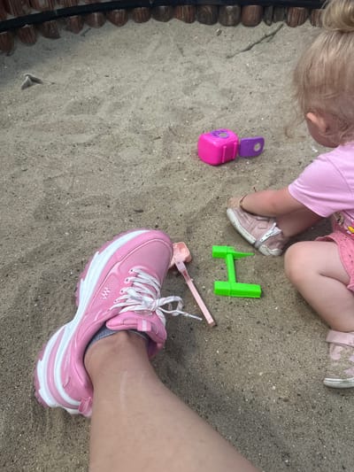 In Donchovci, Bulgaria, a close-up shot captures a sandy area, possibly a playground. In the foreground, a person's left leg is visible, wearing a pink and white athletic shoe. To the right, a toddler, with blonde hair pulled into a bun, sits in a pink shirt and skirt. The toddler's right foot is encased in a pink sneaker, and their left foot is bare. Scattered in the sand are several toys: a bright pink cube with a purple attachment, a pink shovel, and a green toy that resembles an "H" shape. The background features a wooden fence.