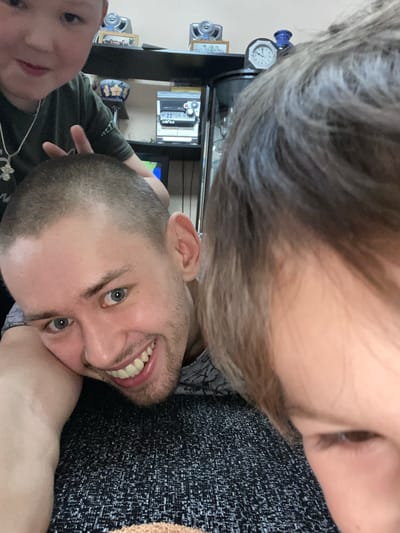 A selfie-style photograph shows three individuals in an indoor setting. A man with short, buzzed hair and blue eyes smiles towards the camera. A child's face is partially visible in the lower left corner. The upper portion of the image shows another person with light-colored hair. They are likely lying on a dark gray, textured surface, possibly a couch. The background reveals shelves with various items, including a clock, speakers, a DVD player, and framed pictures. The location is presumably indoors. There is nothing in the photo that proves it was taken in the city of Bely Klyuch, Russia.