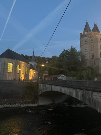 The image depicts a twilight view of the town of Durbuy, Belgium, with the Ourthe River in the foreground. A stone bridge with a curved arch spans the river, carrying a road or walkway across. On the left bank, a stone church with illuminated stained-glass windows is visible, its roof a dark grey. Adjacent to the church, a smaller steeple with a cross rises. To the right, a tall, stone tower with multiple windows and conical roofs dominates the skyline. Trees and foliage cover the hillside behind the buildings. Several streetlights illuminate the scene, casting a warm glow on the church and bridge. The sky is a deep blue, marked by wispy white contrails from aircraft, suggesting a late evening or early morning hour. A dark, thin cable runs diagonally across the upper portion of the image. The river below the bridge is dark and appears to have some ripples or debris on its surface.