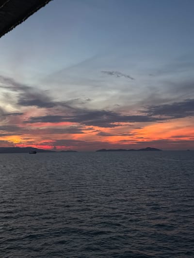 The image depicts a serene seascape at dusk, with the sky ablaze with vibrant colors. A stretch of calm, dark water occupies the lower half of the frame, reflecting the fading light. In the distance, a silhouette of islands lines the horizon, their forms softened by the twilight. A solitary vessel, likely a ship, is visible on the left side of the image, navigating the tranquil sea. The upper portion of the image is dominated by a dramatic sky, transitioning from a pale blue at the top to fiery oranges, pinks, and reds near the horizon, painted with wispy clouds. This view suggests a location along the coast of Thong Nian, Thailand, known for its beautiful sunsets over the sea.