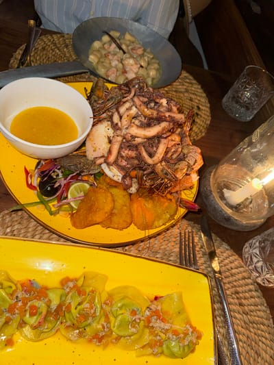The image captures a vibrant, candlelit dinner scene, likely set in a coastal restaurant in Michamvi, Tanzania. The dim, warm lighting, primarily from a single candle encased in a glass bottle, creates an intimate evening atmosphere, enhancing the rich colors of the food.
The dining table is adorned with rustic woven placemats and a lavish spread of food. A large, bright yellow platter dominates the center, piled high with a generous assortment of grilled seafood. This includes numerous succulent octopus or squid tentacles, many large prawns or shrimp, and several pieces of fried plantains or similar fritters. A small, colorful side salad featuring red onion, bell peppers, olives, and a fresh lime wedge complements the seafood. A white bowl of vibrant yellow sauce, possibly a butter or lemon-based seafood dressing, sits alongside.
To the right, another bright yellow rectangular plate presents a row of perfectly arranged pasta. These appear to be large, green tortellini or ravioli, generously topped with a sauce containing diced orange vegetables, likely carrots or squash, and delicate white pieces of what seems to be minced crab meat or other seafood.
On the left side of the table, a smaller, dark metal bowl holds a creamy dish, possibly gnocchi or small dumplings, mixed with pieces of shrimp or white fish in a rich, white sauce. Various clear, textured drinking glasses and silverware are also scattered across the table, ready for use.
Though no people are fully visible, a partial view of a striped fabric, possibly clothing, and a straw hat on the far left suggest diners are present, enjoying this elaborate meal. The extensive seafood selection strongly hints at a location close to the ocean, aligning with the provided context of Michamvi, Tanzania, and suggesting a focus on local, fresh ingredients. The scene evokes a sense of indulgence and a relaxing, enjoyable culinary experience. No text is visible in the image.