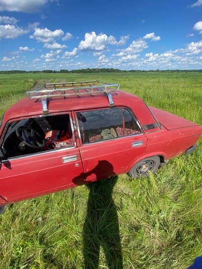 A red sedan with a chrome roof rack is parked on a dirt path in a grassy field under a blue sky with white clouds. The driver's side door of the car is open, revealing the steering wheel, an orange safety vest, and patterned upholstery. The car appears weathered and slightly dusty, with visible rust spots. The field is lush and green, with tall grass extending to the horizon. In the background, trees and a cultivated field are visible. The scene is illuminated by bright sunlight, casting a long shadow from the car and the photographer.