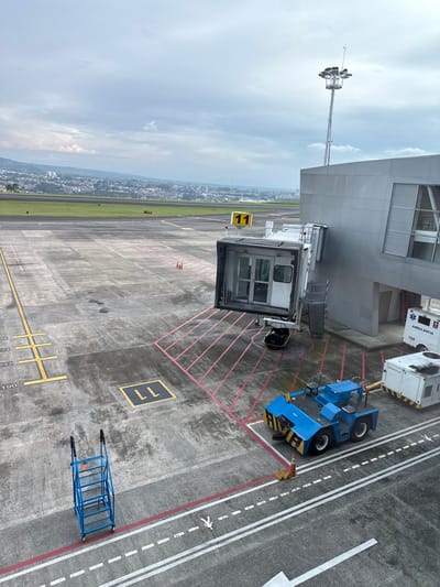 The image captures an airport tarmac and terminal building at an unspecified airport within the city of AMCO, Area Metropolitana Centro Occidente, Colombia, observed from an elevated vantage point under a cloudy sky.
The foreground and midground consist of a grey concrete apron with yellow, white, and red markings, including the numbers "190," "232," "272," and "737" along a yellow line, and a square "LL" marking. A yellow sign displaying "11" is positioned centrally. A white and grey jet bridge, designated Gate 11, extends from a grey, multi-story terminal building on the right. The jet bridge's forward section is open and not connected to an aircraft. Red diagonal lines are painted on the apron surface beneath the jet bridge, indicating a parking zone.
Adjacent to the terminal building and beneath the jet bridge, various ground support equipment is present. This includes a blue aircraft tug/pushback tractor, partially obscured by an orange traffic cone, with "LEYBLAN" visible on its side. Behind the tug, a white ambulance vehicle marked "AMBULANCIA" is parked, alongside a white rectangular service cart. A tall metal light tower with multiple floodlights extends above the terminal building's roofline.
Further to the left on the apron, a blue, four-step rolling ladder is positioned. In the background, a green grass strip separates the apron from a distant runway or taxiway. Beyond this, a sprawling city with numerous buildings is visible, set against a backdrop of hills or mountains. No people are depicted within the scene.