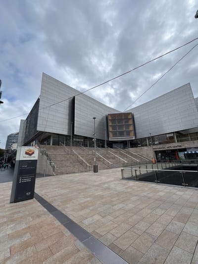 The image shows the exterior of the Performing Arts Centre in Roseville Chase, Australia, under a cloudy sky. The modern building features a silver, angular facade. In the foreground, there is a paved plaza with a dark grey directional sign for "THE CONCOURSE" which lists "Concert Hall", "Theatre", "Lounge", and "Chatswood Library" as destinations. Below these are rules stating "Cyclists Must Dismount", "No Skateboards", and "No Dogs Off leash". A wide set of stone stairs leads up to the building's entrance. A glass and metal railing runs along the edge of a lower level to the right of the stairs. Some people are visible walking in the background, and the facade of another building can be seen to the left. Thin wires are visible strung across the sky in the upper portion of the image.