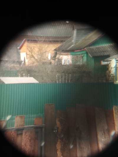 The image, possibly taken through a monocular or similar optical device, shows a view across a fence in Высокиничи, Russia. In the foreground, a dilapidated wooden fence of varying heights and weathered planks is visible. Behind this, a solid green metal fence extends horizontally across the middle ground. Beyond the green fence, a blurred residential landscape features buildings with pitched roofs covered with gray tiling. One building appears to be painted brown, and another is green with red trim. Bare trees are interspersed between the buildings. The sky is obscured by the roofs and the optical device's vignetting. A satellite dish is visible on the roof of the green building.