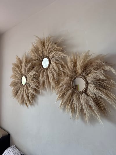 In Kosharitsa, Bulgaria, a beige wall serves as the backdrop for a trio of decorative mirrors. Each mirror is encircled by a halo of dried pampas grass, creating a sunburst effect. The mirrors themselves are small and circular, with visible reflections of the surrounding room, including a glimpse of a framed picture on one of them. The pampas grass is a light, earthy tone, varying in shades of beige and light brown. The mirrors are arranged horizontally across the wall, with slight variations in their placement and the density of the surrounding grass. The lighting appears natural, casting subtle shadows on the wall. The overall aesthetic suggests a bohemian or naturalistic interior design.