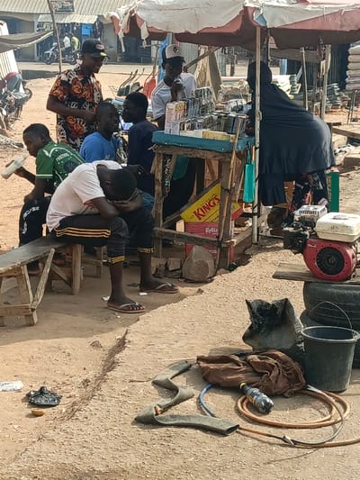 A group of people are gathered near a market stall under an umbrella. A man sits on a bench with his head in his hands. A woman dressed in black stands behind the stall. In front of the stall, there is an engine, tires, a bucket, a torn inner tube, and a water bottle on the ground. Other people are visible in the background.
