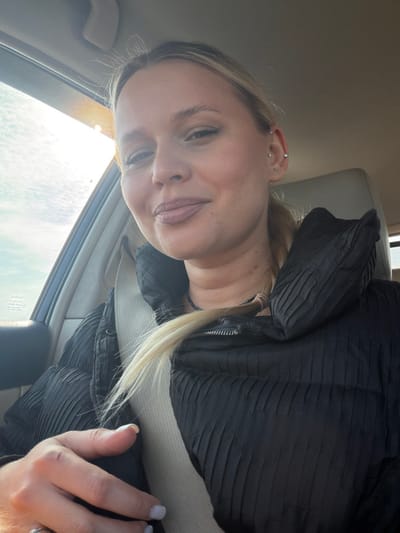 A young adult female is depicted in a selfie taken inside a vehicle in Povarenki, Russia. She has light skin, blonde hair styled back, and multiple ear piercings on her left ear. She is wearing a dark, possibly black, horizontally ribbed or quilted puffer jacket and has a light-colored seatbelt diagonally across her chest. Her left hand, with light-colored nail polish, rests near the lower part of the seatbelt. The woman is looking directly at the camera with a slight smile. Through the window on the left side of the vehicle, bright sunlight is visible against a partly cloudy sky, indicating daytime. Faint text, possibly "TOYOTA," is reflected on the window. The interior of the vehicle appears to be light-colored.