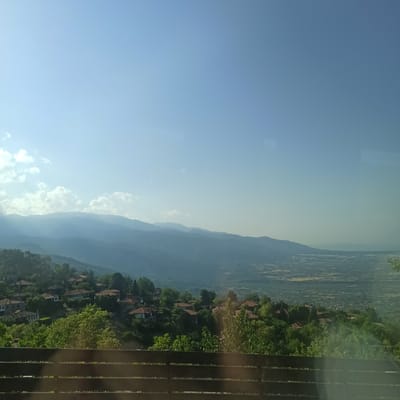This is a vertical, eye-level, medium-shot photograph taken from a high vantage point. The scene is a sprawling landscape with layers of mountains receding into the distance under a clear blue sky. The sky is bright and appears to be from the daytime, possibly morning or afternoon, with minimal cloud cover, save for a few wispy clouds visible near the horizon. The light suggests a sunny day.

In the foreground, to the right, there is a wooden railing, which appears to be part of a balcony or viewing platform. The railing consists of dark brown, vertical slats. Beyond the railing, the landscape unfolds. A village or settlement is nestled into the hillside, characterized by numerous houses with terracotta tile roofs, interspersed with dense green trees. The houses are scattered and follow the contours of the terrain, creating a picturesque view. The vegetation is lush and varied, with a mix of trees and shrubs.

The background is dominated by undulating mountain ranges that fade into a hazy blue due to atmospheric perspective. The distant valley floor is visible between the mountain ranges, appearing as a patchwork of fields or possibly another settlement.

The overall mood is serene and expansive, offering a breathtaking view of a rural or semi-rural mountainous region. There is no visible text in the image. The image appears to be a landscape shot, potentially taken from a tourist viewpoint or a residential property with a scenic overlook, possibly in Neos Panteleimonas, Greece, given the context.