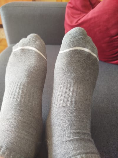 This is a close-up, low-angle shot of two feet wearing grey ankle socks with a white stripe near the top cuff. The socks have a ribbed texture, and there are some faint indentations on the fabric, suggesting they have been worn. The feet are resting on a dark grey textured surface, likely a couch or armchair. In the background, out of focus, is a bright red cushion and a hint of a light-colored wooden floor. The lighting appears to be soft and natural, suggesting it's daytime. The scene evokes a relaxed, comfortable atmosphere, possibly indicating a moment of downtime. No visible text is present in the image. The location context is Cēsis, Latvia.