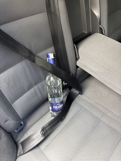 The image shows a bottle of Finlandia vodka secured by a seatbelt on the back seat of a car. There are no people visible. The car seat is gray with a textured surface. The bottle is approximately half full of a clear liquid. The environment is limited to the car's interior. There is no evidence of the location being in Dolno Drianovo, Bulgaria, or any other specific location.