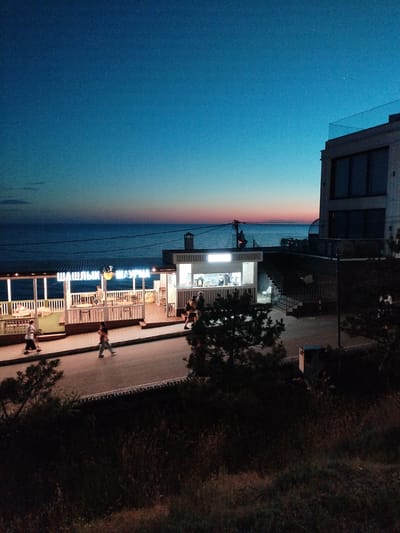 At dusk, a coastal promenade in Vinogradny, Russia, features a dining establishment with illuminated signage that reads "ШАШЛЫК ШАУРМА" (Shashlik Shawarma). The open-air seating area of the restaurant is visible, with tables and chairs set up, and several people are seated inside. In front of the restaurant, a paved walkway runs parallel to the sea, and a few individuals are walking along it. Beyond the walkway, the sea stretches to the horizon, where the sky displays a gradient of deep blue to orange and pink hues from the setting sun. To the right of the promenade, a modern multi-story building stands, its upper floors illuminated, and a glass railing adorns the rooftop. The foreground is dominated by dark, out-of-focus vegetation and a road.