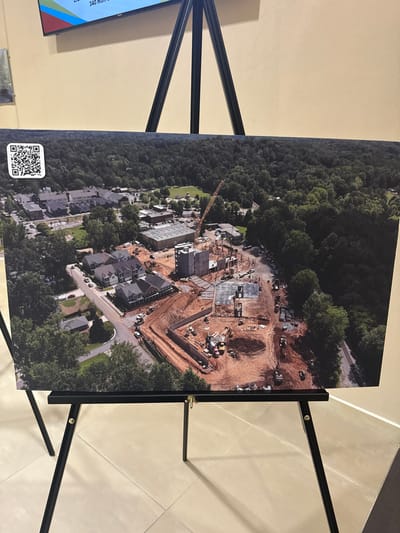 An aerial photograph depicts a construction site adjacent to a densely wooded area. A large, angular excavation forms the rough shape of a 'V', indicating potential underground construction or foundation work. Alongside, building foundations and some partially constructed structures are visible. Existing residential housing occupies the lower part of the photo, with trees interspersed between the houses and the ongoing construction. A yellow crane is also present on the site. A QR code is in the lower left.