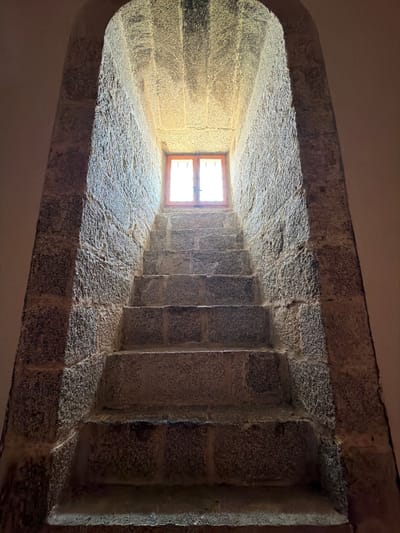 This is a low-angle, full shot of a stone staircase leading up to a small, arched window. The staircase is made of rough-hewn, grey granite blocks, with seven visible steps. The walls surrounding the staircase are also made of similar stone and curve inward to form an arched opening. Sunlight streams through the window, casting a warm glow on the upper portion of the stairs and the ceiling of the arch. The window itself appears to be a simple wooden frame with glass panes, divided into four sections. The environment is dimly lit, suggesting an interior space within a historical building. The image was taken in San Lorenzo De El Escorial, Spain, likely within the Monastery of San Lorenzo de El Escorial, given its architectural style and location. There are no people or animals present in the image.