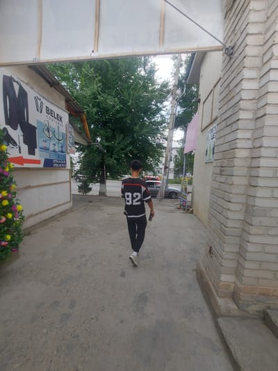 A young man wearing a black and white jersey with the number 92 on the back walks away from the camera down a concrete walkway. He is wearing black pants and white socks with sneakers. He is holding a black object in his right hand. The walkway is flanked by a brick building on the right and a wall with a sign on the left. The sign on the left has images of suits and formal wear, and text in a foreign language. A decorative bush with yellow and purple flowers is on the far left of the image. The background shows trees and buildings, suggesting an urban environment. The scene appears to be outdoors, under an awning or covered walkway. The sky is visible above the trees. The overall lighting suggests daytime, possibly overcast.