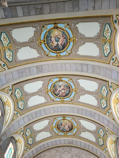 The image shows the interior ceiling of a building, likely a church, with ornate painted details. The ceiling is arched and decorated with multiple circular medallions containing paintings of cherubic figures. These medallions are set within intricate gold and beige borders with green accents. Below the medallions, there are rectangular panels with decorative white scrollwork. The ceiling is constructed from stone blocks interspersed with painted plaster. Stained glass windows are partially visible on the left side of the image, indicating a religious setting. The scene appears to be in Terras de Bouro, Portugal, suggested by the architectural style often found in historical Portuguese buildings.