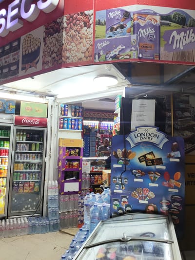 The image shows the exterior of a small convenience store, likely in an urban area. The store's facade is adorned with colorful advertisements for various products, including chocolates, nuts, and soft drinks. Several refrigerated display cases are visible, stocked with beverages and other items. A sign advertising "London Dairy Premium Ice Cream" is prominently displayed, featuring images of different ice cream products. Bottles of water are stacked on the floor outside the store. A man can be seen standing inside the store, partially obscured by a display. The overall impression is that of a bustling local shop with a wide selection of goods.