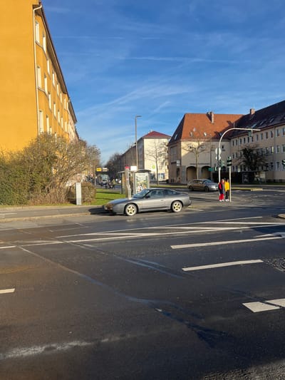 The image presents an urban street scene in Schweinfurt, Germany, under bright, clear daylight. The perspective is slightly elevated, overlooking a multi-lane road.

In the immediate foreground, a light gray two-door sedan is parked or stopped at the edge of the asphalt road, facing away from the viewer. The road is marked with clear white lines, including lane dividers and possibly directional indicators. Adjacent to the road, a narrow grassy median with several bare-branched trees separates the roadway from buildings.

In the midground, a pedestrian wearing a bright yellow jacket and red pants stands near a traffic light pole on the sidewalk, appearing to wait to cross the street. A darker colored car is stopped behind them. An information pillar or bus stop sign with unreadable advertisements is also visible. Beyond the median, a row of multi-story buildings lines the street. One building features a red-tiled roof and a brown facade. On a lighter-colored building further in the background, the word "Kindergarten" is partially visible.

To the far left of the frame, a large yellow building with numerous windows occupies a significant portion of the view. The sky above is a vibrant blue with a few faint white contrails, indicating sunny and fair weather. The trees' bare branches suggest the season is likely late autumn, winter, or early spring. The scene depicts a typical, calm urban environment with traffic and pedestrians.