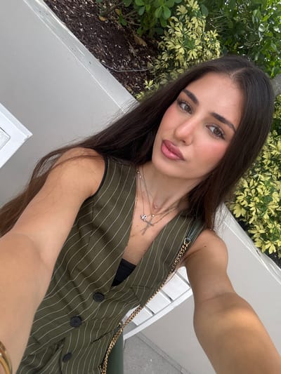 A young woman with long dark hair is taking a selfie outdoors. She is wearing a green pinstripe vest over a black top and several necklaces. She has light skin and is wearing makeup, including lipstick. Her arm is extended, holding the camera. Behind her, there is a light-colored bench, foliage with light green and yellow leaves, and dry ground cover. The setting appears to be a public outdoor space in Hallandale Beach, Florida, United States of America, possibly a park or garden area.