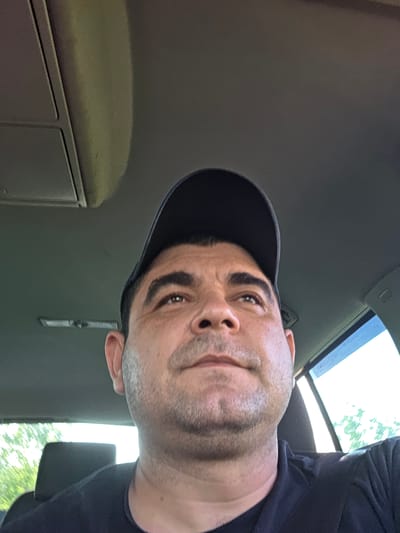 A man wearing a dark baseball cap and a dark shirt is sitting in a car. The interior of the car is dark grey or black. A window is visible in the lower left corner, showing trees and sky outside.