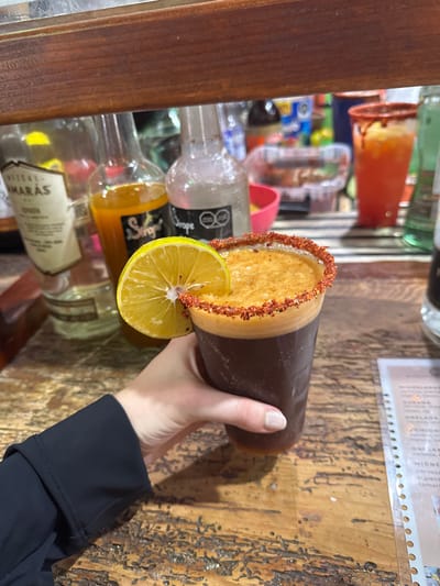 The image presents a close-up, eye-level view of a beverage held within an establishment in Huasca de Ocampo, Mexico. A left hand, featuring lightly polished fingernails and clad in a dark, long-sleeved garment, grasps a transparent plastic cup containing a dark brown, foamy beverage. The cup's rim is thickly coated with a reddish-orange spice, and a wedge of fresh lime is affixed to the rim. The hand and cup rest on a rustic wooden table with distinct grain and dark, textured imperfections. In the background, behind a horizontal wooden beam, multiple bottles are visible, including one labeled "AMARAS" (likely Mezcal Amarás), an amber liquid labeled "Sirope," and a clear liquid also labeled "Sirope." Further back, a red plastic cup with a spiced rim and a reddish-orange liquid is partially visible, along with other bar essentials. A laminated menu detailing various drink options, including "MICHELADA" and "CERVEZA," is partially visible on the table to the right.