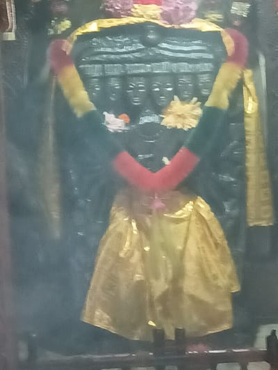 The image shows a close-up of a decorated statue, likely of a deity, within a temple in Gojra, India. The statue is adorned with garlands of red and yellow cloth, and a shimmering golden fabric is draped around its lower half, resembling a skirt. A yellow floral ornament is placed on the statue's chest. The background appears to be a dark, carved stone structure, possibly part of the temple's architecture. The overall lighting is dim, with some areas illuminated by what appears to be artificial light, creating a hazy effect. The image's resolution is low, with a grainy texture and blurred details.