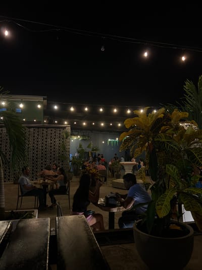 This image depicts a nighttime scene at an outdoor dining or social venue located in Santiago, Panama. The area is illuminated primarily by multiple strings of warm-toned incandescent bulb lights strung overhead against a dark night sky. Numerous patrons are seated at various tables on a paved surface.
In the midground, to the left, a man in a light short-sleeved shirt is seated at a table, facing right. To the center-right, a woman with long dark hair, wearing a dark top and light-colored shorts, is seated at a small wooden table, facing left. Across from her, a man in a light blue short-sleeved shirt is seated, observed looking down at a dark object on the table, possibly a mobile phone. A dark object, potentially a condiment holder, is also present on their table.
Multiple other individuals and groups are visible seated or standing throughout the background, occupying various tables. Large potted plants with green and yellow variegated foliage are interspersed within the seating area, including a prominent specimen in the immediate foreground on the right. A building with a light green upper section and a textured, patterned lower wall, possibly concrete block, is visible on the left side of the frame. An indistinct dark structure partially obscures the immediate bottom foreground.