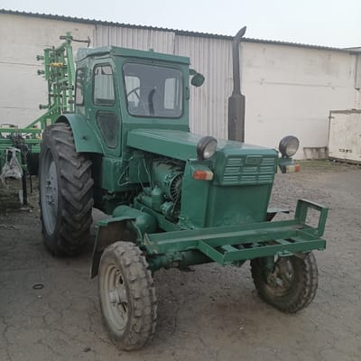 This media file features a green tractor parked outdoors. The tractor is the central object, with its large rear tires and smaller front tires visible. It appears to be a utilitarian model, possibly used for agricultural purposes, as evidenced by the attachment mounted on the rear, which resembles agricultural machinery. The tractor has a cab with windows and a visible steering wheel inside. A tall exhaust pipe rises vertically from the engine area.
The setting is an outdoor area with a rough, dirt or gravel ground. In the background, a plain, white building with a corrugated metal roof runs horizontally across the frame. The sky is overcast, suggesting a cloudy or hazy day, possibly early morning or late afternoon due to the diffused light. There are no people visible in the image. The overall impression is one of a functional vehicle in a utilitarian or rural environment. No text is discernible in the image.