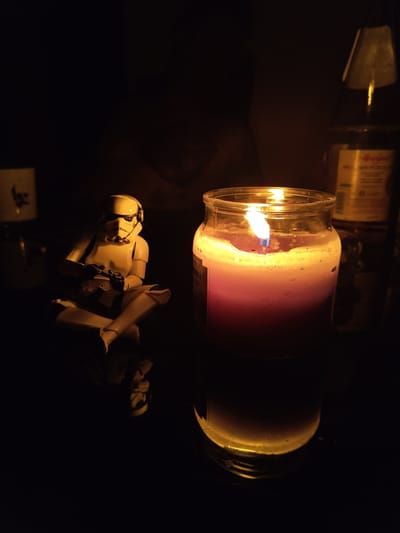 The image shows a horizontal, lit candle in a glass jar, with a Star Wars stormtrooper action figure reclining beneath it. The lighting is dim, primarily illuminated by the candle, casting shadows and creating a dark environment. There are other objects visible in the background, but they are largely obscured by the darkness. The setting and location (Schmelz, Germany) cannot be ascertained from this image alone.
