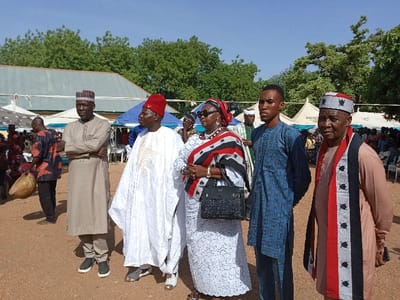 The image shows six people standing in a dirt lot in front of a building and some tents, all wearing traditional Nigerian clothing. The location is Zum, Nigeria at the coordinates (latitude: 10.116, longitude: 13.046). They are all looking in the direction of the camera. The person on the left is holding his hands crossed in front of him and is wearing a long, beige robe. The person on the right is wearing a long tan robe and a red and white patterned scarf. The three people in the middle are wearing long robes and scarves of various colors. The person on the left of the group is wearing a long white robe with a red hat and carrying a white staff. The person in the middle is wearing a white dress and a red and black scarf, and she is holding a black handbag. The person on the right of the group is wearing a long blue robe. There are many trees in the background, as well as a building and tents. There are other people in the background. The weather is sunny. The ground is dirt.
