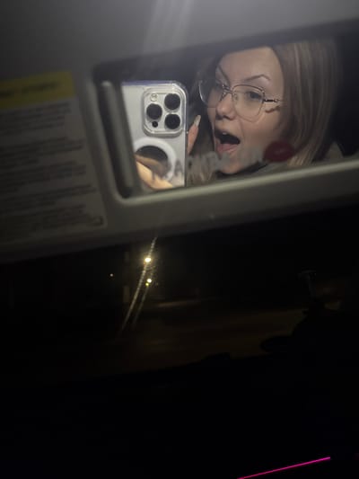 The image displays a selfie taken at night from inside a vehicle, captured as a reflection in an overhead vanity mirror. The mirror, housed within a light-colored plastic frame, shows the upper torso of a fair-skinned female individual. She has light-toned hair and wears eyeglasses with clear frames. Her mouth is open, revealing teeth, and her expression is animated. A prominent red lip-shaped print is visible on the mirror's surface near her cheek. Her left hand, with long, light-colored manicured fingernails, holds a light-colored smartphone with a clear case and a triple-lens camera array, positioned to capture the reflection. A circular magnetic accessory is visible on the phone case. To the immediate left of the mirror, a portion of the car's sun visor with a yellow warning label is visible. The lower section of the image shows the view through the vehicle's windshield at night, revealing a dark environment with several distant, glowing circular lights, consistent with streetlights or vehicle lights on a road. Faint streaks or smudges are present on the windshield. A thin, horizontal glowing pink light line extends across the lower right corner, indicating internal ambient lighting. No specific landmarks or identifying features are present to determine a precise location within the city of Skalsko, Bulgaria.