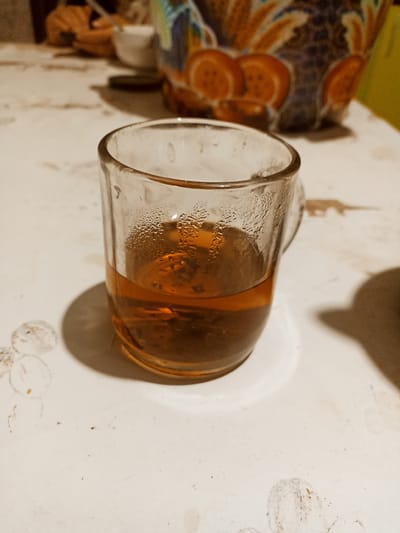 This image captures a close-up view of a glass mug filled with a dark amber liquid, likely tea, resting on a white, slightly textured surface. Condensation is visible on the exterior of the glass, suggesting the beverage is hot. The mug has a handle, though only a portion of it is visible.
In the background, out of focus, are hints of a table setting. To the left, there's a suggestion of people's hands and possibly other dishes. To the right, a vibrantly colored, patterned object, possibly a decorative bowl or vase with orange and blue motifs, adds a splash of color to the scene. The overall lighting suggests an indoor setting, possibly in the evening or a dimly lit room. The surface appears somewhat worn, with faint smudges or stains visible, giving a lived-in feel.
The image is taken from a slightly elevated angle, focusing primarily on the mug of tea. The scene appears to be set in a casual, domestic environment. There is no visible text in the image. The location context is Кош-Дөбө, Kyrgyzstan.