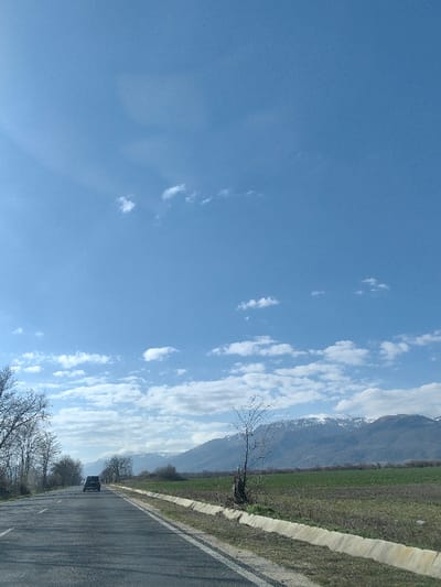The scene depicts a rural road environment in the vicinity of Dolno Sahrane, Bulgaria, under a clear blue sky with scattered white cumulus clouds. A two-lane asphalt road extends into the distance, showing a solid white line marking its right edge. A dark-colored sport utility vehicle (SUV) is observed in the mid-ground, traveling away from the viewer on the right side of the roadway. To the left of the road, a line of dormant deciduous trees with bare branches runs parallel. To the right of the asphalt, a concrete drainage ditch parallels the road, beyond which lies an expanse of cultivated green field. A single, bare deciduous tree with a short stump is positioned within this field, adjacent to the ditch. In the distant background, a range of mountains with visible snow caps spans the horizon. No individuals are present in the frame.