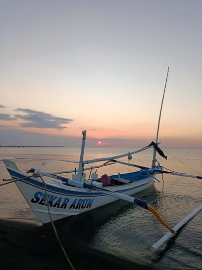 A fishing boat named "SEKAR ARUM" is moored at the shore in Jangkar, Indonesia. The boat is white with blue trim and lettering. It has two outriggers extending from its sides. The sun is setting over the calm sea, casting an orange and pink glow across the sky and water. The shore is dark sand. In the distance, a long pier stretches out into the water, with a few buildings visible on the horizon. The overall scene depicts a peaceful end-of-day in a coastal Indonesian town.