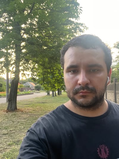 A man with a beard and mustache, wearing a black t-shirt and white earbuds, takes a selfie outdoors. Behind him, a tree stands prominently to his left, with sunlight filtering through its leaves. Further in the background, a road runs parallel to a row of trees, and beyond that, houses and fences are visible. The overall setting appears to be a residential area in the city of Nikopol, Ukraine.