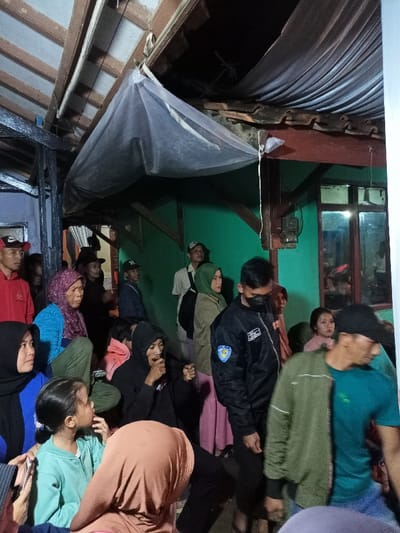 A diverse group of people, comprising men, women, and children of various ages, are gathered in what appears to be a communal, sheltered space in Pangalengan, Indonesia. The setting features exposed wooden beams supporting a roof structure, with a white, translucent fabric (possibly a mosquito net or decorative cloth) draped across the ceiling. A vibrant green wall with a window is visible in the background, along with a brown wooden post holding an electrical meter.
The individuals are either standing or sitting on the ground, most of them looking in different directions, suggesting they are observing an event or conversing. A young girl in a light green top looks upwards with a curious expression. Many adults wear casual clothing, including t-shirts, hoodies, and hijabs. One man in the center wears a black jacket with a shield-shaped badge on the arm and a black face mask, while another in the foreground wears a blue t-shirt and a dark baseball cap.
The lighting is dim, indicating it is likely evening or nighttime, with artificial light sources illuminating the scene, casting soft shadows. The overall atmosphere appears calm, with no strong emotions visibly expressed by the crowd. There is no clearly discernible text in the image.