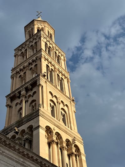 This is a low-angle, upward view of the Bell Tower of Saint Domnius Cathedral in Split, Croatia, against a cloudy sky. The tower is made of light-colored stone and features intricate architectural details, including arched windows, columns, and decorative carvings. The perspective emphasizes the tower's height and grandeur. The sky is a mixture of blue and gray clouds, suggesting it may be a partly cloudy day. The lighting indicates it is daytime, with the sun casting shadows on the stone. There are no people visible in the image. The overall mood is one of historical monumentality and architectural beauty.