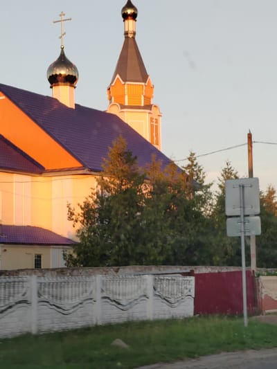 This image shows a large building with a purple roof and orange accents, likely a church, with a prominent dome topped with a cross. Adjacent to it, another tower rises, also with orange detailing and a dark spire. Evergreen trees are visible behind the building and in front of the fence. A decorative white fence with dark wavy patterns runs across the foreground, separating the church area from a grassy area and a road. A red gate is visible to the right of the white fence. A pole with two square signs is also present on the right side of the image. The lighting suggests it is either early morning or late afternoon, with warm sunlight casting a glow on the building. The sky is clear and pale blue, indicating fair weather. No people are visible in the image. The scene is likely set in a rural or semi-rural area, given the presence of the fence, grass, and trees. No text is discernible in the image.