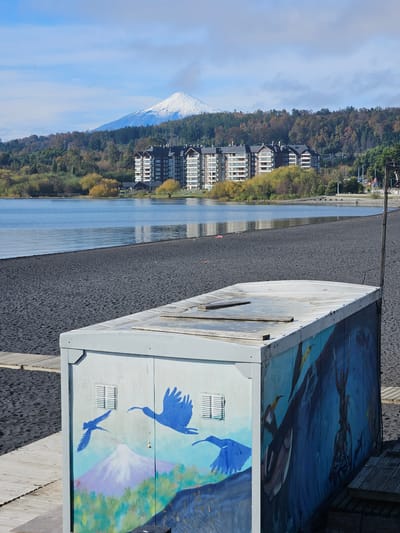 The image depicts a scene in Villarrica, Chile.  A painted electrical box stands in the foreground, featuring artwork of birds and an underwater scene. Beyond the box is a dark-colored beach running along the edge of a body of water.  Buildings are clustered along the shoreline, backed by dense forest.  In the distance, a snow-capped volcano is visible under a blue sky with scattered clouds.