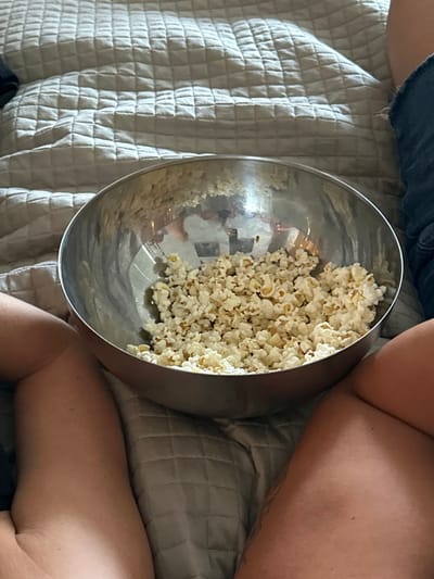 An overhead, first-person perspective image captures a scene within the Merom HaGalil Regional Council, Israel. The viewer's legs are visible, positioned symmetrically in the lower half of the frame, with a large, silver-toned metallic bowl filled with popped popcorn resting between them on a light grey, quilted bedspread. The individual's left leg is bare, while the right leg is partially covered by dark blue denim fabric, likely jeans. The popcorn in the bowl is white and yellow-tinted, consistent with a buttered or seasoned variety. Reflections on the bowl's polished surface show distorted views of the upper environment, including what appears to be ambient room lighting. The bedspread exhibits a consistent square quilting pattern and minor creasing.