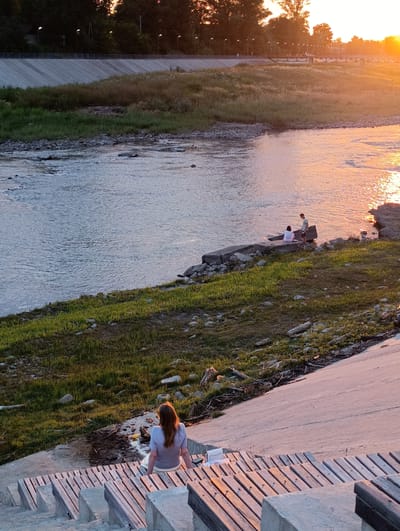In the city of Kelermesskaya, Russia, a serene riverside scene unfolds at sunset. The sun casts a warm, golden glow across the water, reflecting on its rippling surface. On the bank, a young woman with auburn hair sits on wooden stairs, facing away from the camera, her gaze directed towards the tranquil river. Further along the embankment, two figures, a woman and a man, are seated on a concrete ledge, their backs to the viewer, enjoying the picturesque view. The river is bordered by a grassy slope and a concrete embankment, with trees and a bridge visible in the distance under the soft evening light.