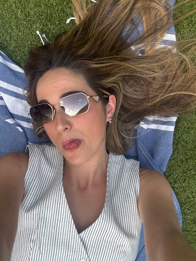 The image shows a young woman lying on her back on a blue and white striped towel on a grassy field. She is wearing a white sleeveless top with thin blue stripes and large, square-shaped sunglasses with a golden frame. Her hair is long and blonde and is partially covering her face. The woman is looking directly at the camera and sticking her tongue out. The image appears to be a selfie taken from a low angle, as the woman is lying down and the camera is positioned above her. The location of the photo is a grassy field in Majadahonda, Spain. The coordinates provided (40.469, -3.857) pinpoint the location to be a residential neighborhood with green spaces. The woman appears to be enjoying a relaxing afternoon in the park.