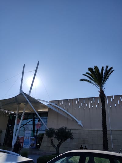 A bright, sunny day in Cebalat, Tunisia, is depicted in this vertical, medium-angle shot. The clear blue sky dominates the upper half of the frame. In the mid-ground, a modern building with a white, tensile fabric roof structure is visible. The sun is positioned directly behind the roof's supports, creating a lens flare. The building's facade features large glass windows adorned with decorative floral images and signage in Arabic. To the right, a tall palm tree stands against a concrete wall with a decorative pattern of small, rectangular cutouts. In the foreground, the hoods of two parked cars are visible. A man in a dark shirt stands near the entrance of the building on the left. Two figures are silhouetted against the wall on the right, near the palm tree. A small olive tree is situated between the building and the palm tree. The overall scene suggests an outdoor commercial or public space.