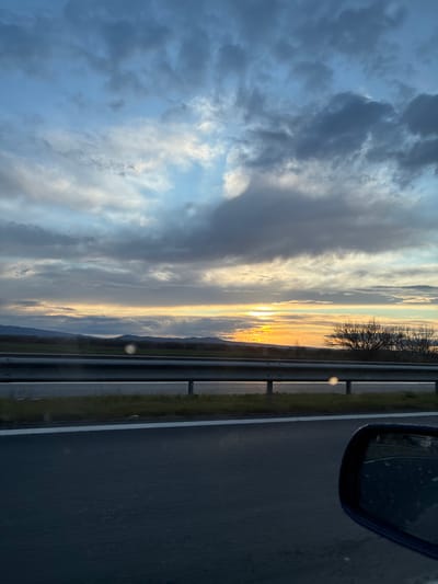 The image shows a view from a vehicle traveling on a road. A side mirror of the vehicle is visible in the upper right corner. A highway barrier runs parallel to the road. The horizon is visible in the distance with mountains. The sky is filled with clouds and the sun is setting. There is a silhouette of trees near the horizon. The location could not be determined based on the data provided.