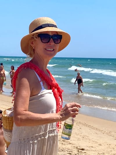 An eye-level medium shot captures a smiling Caucasian woman in her 50s at the beach in Conil de la Frontera, Spain. She wears a straw hat with a black band, large dark sunglasses, a coral-red scarf draped over her shoulders, and a light-colored, sleeveless dress with intricate lace detailing. She is holding a clear plastic bottle of water with a green label in her right hand, which is extended forward. Her left arm is bent, and she is looking to her right, towards the sea.
The beach is sandy, with footprints visible. In the background, the turquoise ocean has moderate waves, and several people are visible swimming and walking in the water. To the left of the main subject, two other women in swimwear are standing on the wet sand closer to the water. Further left, a man's face is partially visible in the foreground, looking left. The sky is clear and blue. The overall scene suggests a sunny day at the beach.
